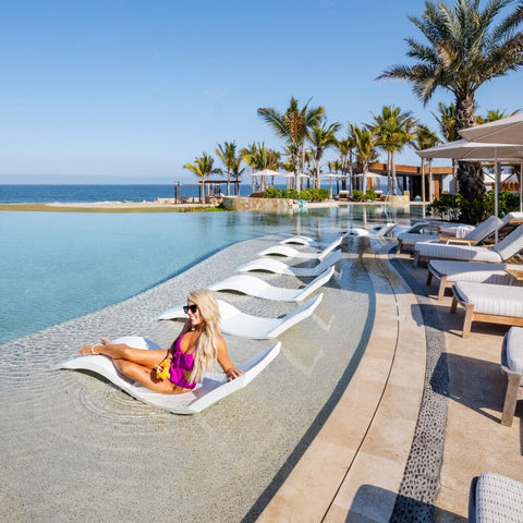 Woman lounging on white Ledge Lounger in-pool chaise in a row of white in-pool chaises on ledge