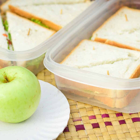 Plastic containers with two meat and cheese sandwiches cut into triangles, with a green apple on a plate