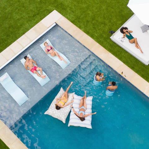 Overhead shot of pool surrounded by grass and people on Ledge Lounger in-pool chaises, Laze pillows, and barstools, and a person laying on a daybed.