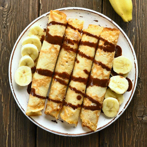 Plate of four rolled up crepes drizzled with chocolate sauce, with sliced bananas on either side