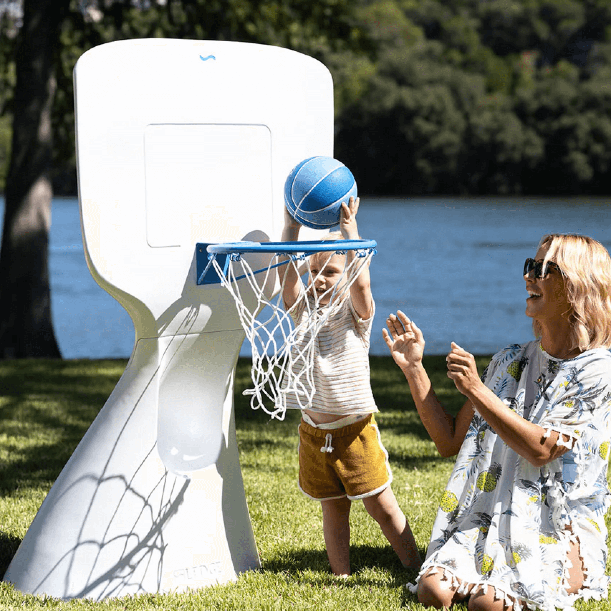 Child playing basketball with woman by a lake.