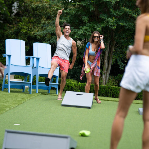 Couple playing cornhole and man cheering, with two Mainstay Adirondack Tall chairs behind them