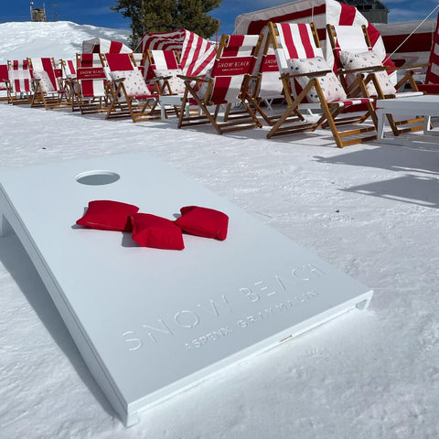 White cornhole with red bean bags on snow, with striped chairs in background