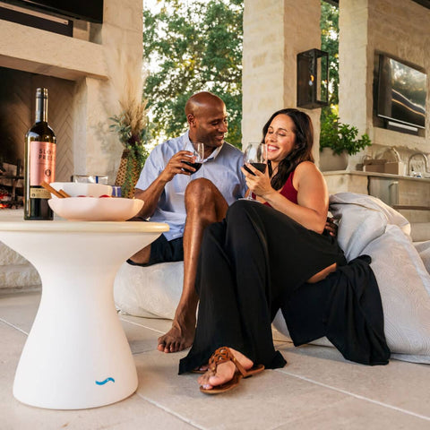 Couple lounging on cushions and drinking wine on backyard patio, with wine and snacks on a side table