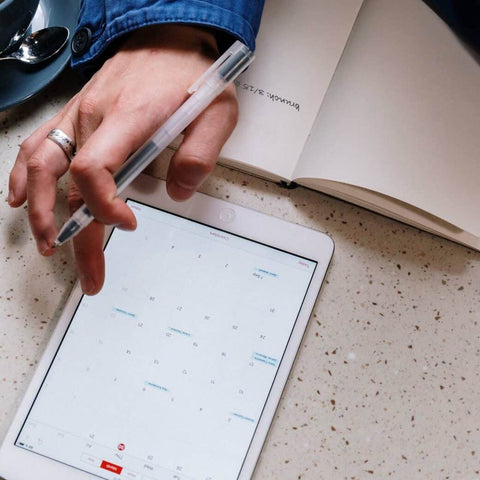 Hand with pen hovering over tablet with calendar on it, and open notebook next to it