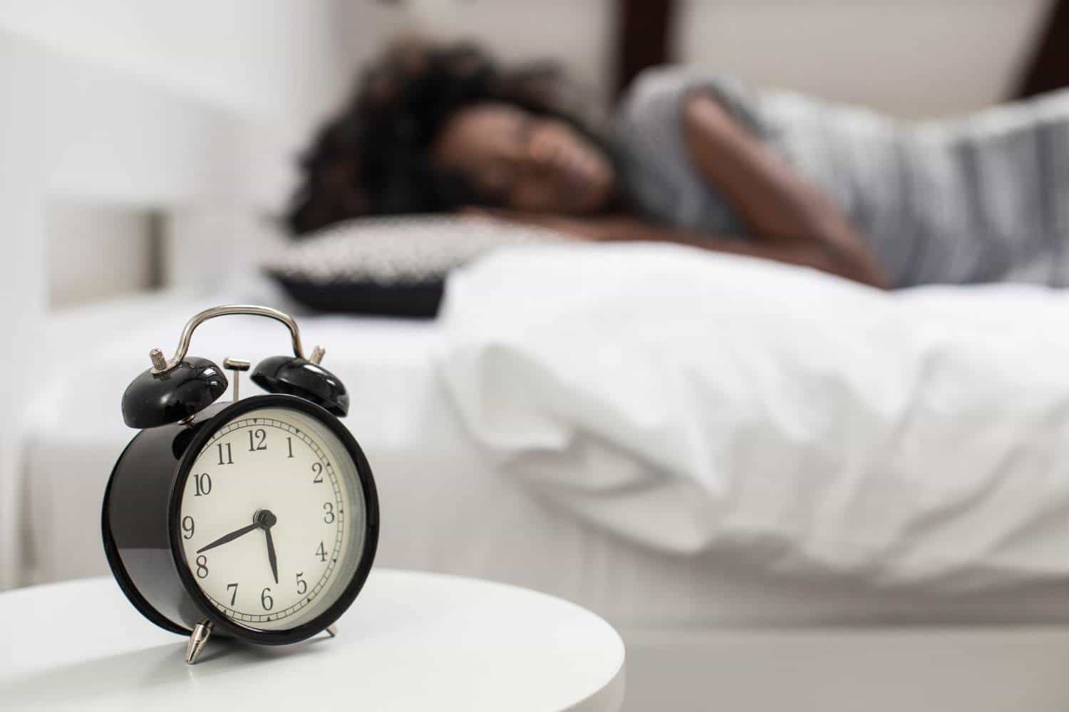 A woman is laying in bed next to an alarm clock.