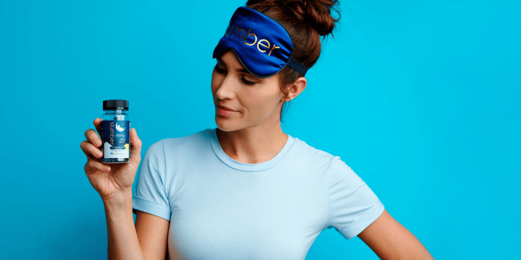 Woman wearing a 'Slumber' branded sleep mask, holding a bottle of Slumber sleep aid capsules, against a blue background, promoting restful sleep.