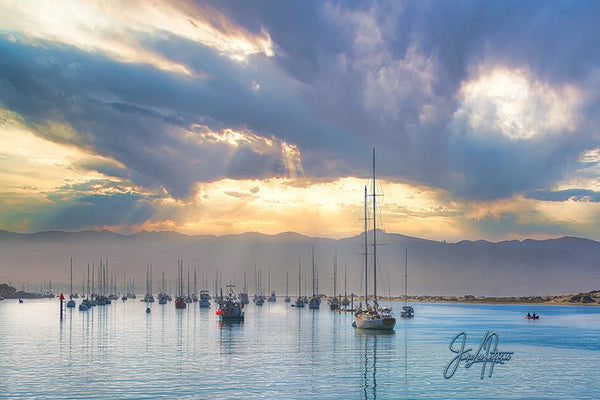 The calm waters of Morro Bay cradle a fleet of sailboats under a dramatic sunset sky, where rays of light break through the clouds, highlighting the serene and picturesque maritime scene.