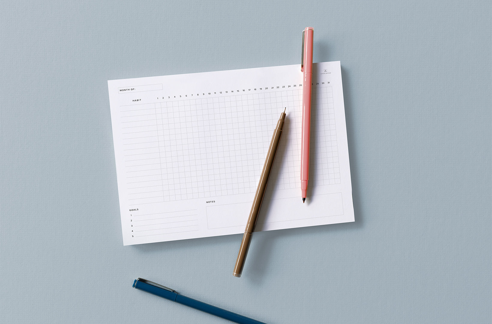 The Appointed Habit Tracker pad sits against a light gray background. Three fine-tip Le Pens in various colors are strewn across the surface.
