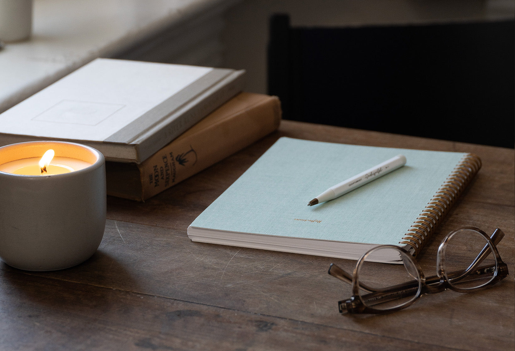 The Reflections Journal sits on a cozy wooden desk. A lit candle and other accessories including glasses and a pen sit aside. Light streams through a window.