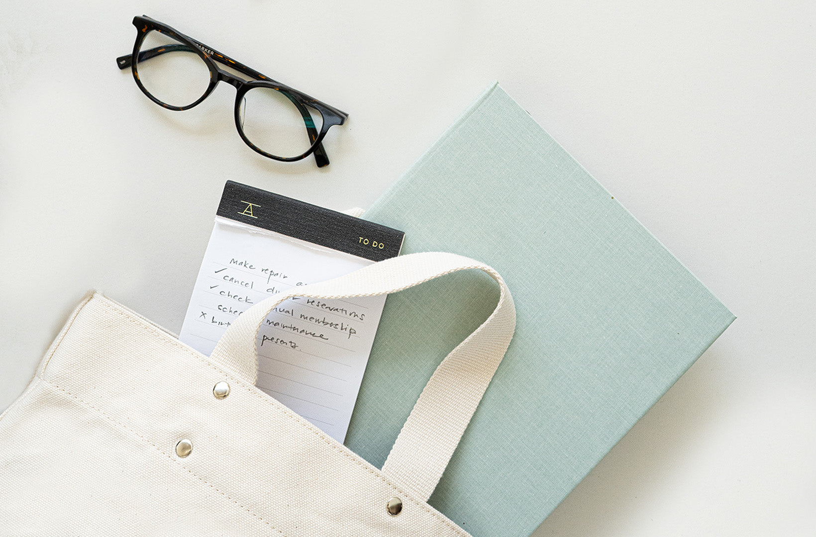 A canvas tote bag sits against a light gray background with contents spilling out, including the 23-24 compact binder planner, to do pad, and glasses