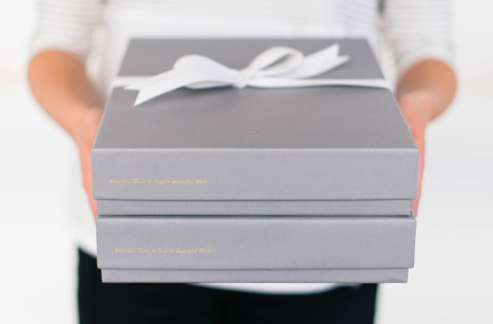 Hands reach out to hand you a dove gray gift box wrapped in white satin ribbon