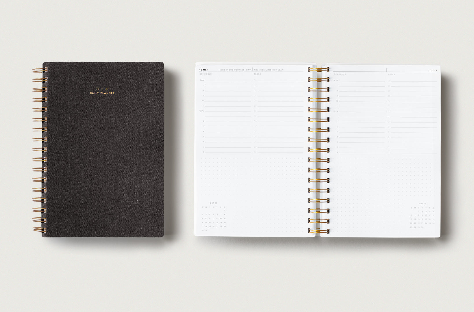 Two new 22-23 Wire Daily Planners sit on top of a light beige background. One is closed showing the textured Charcoal Gray bookcloth, and one lies open displaying the hourly daily format with room for notes