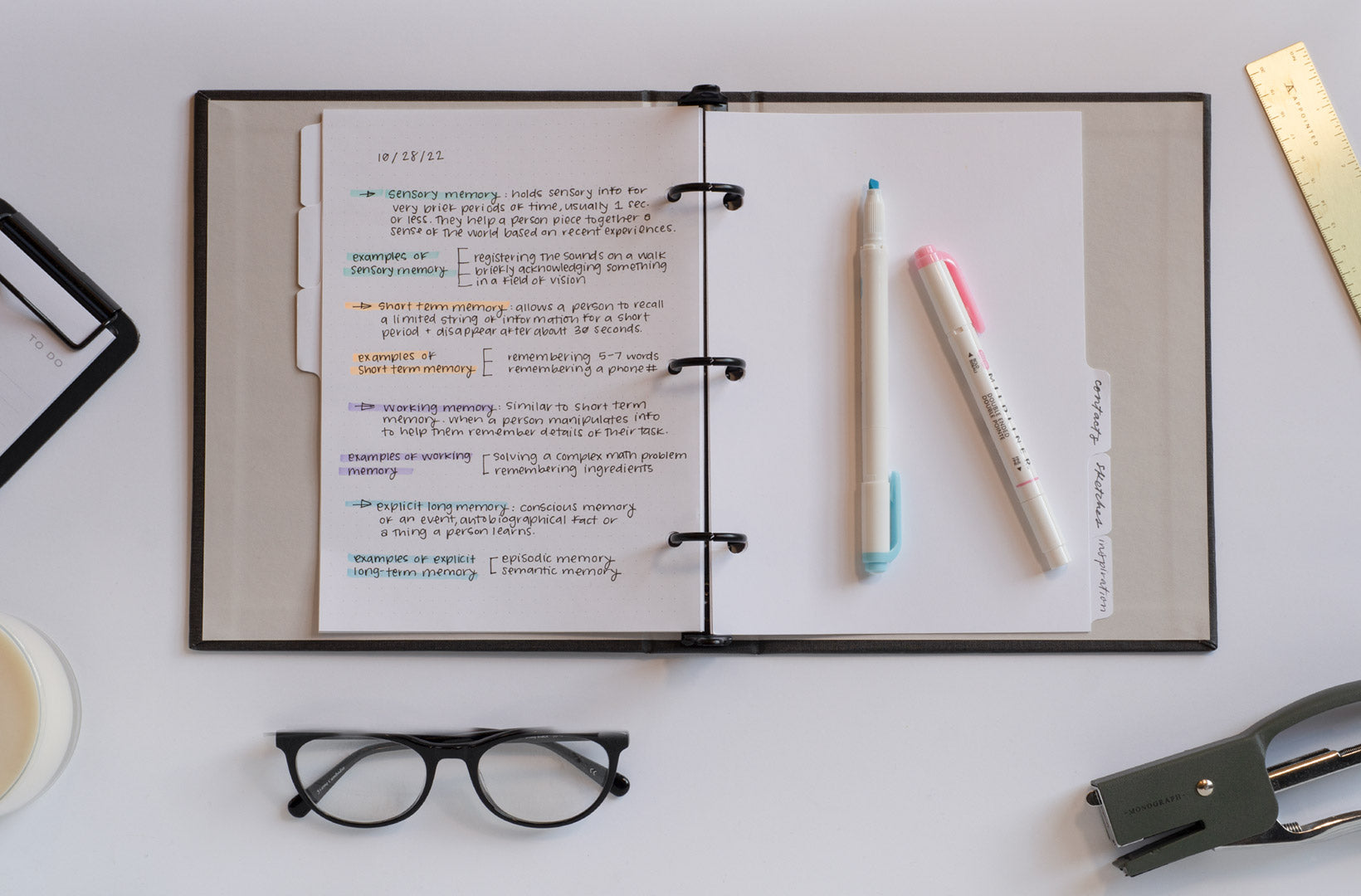The Compact Binder lies open on a white desktop. Class notes are filled in on the left-hand side of the lined pages. Highlighters sit on top of the planner and other accessories are strewn to the side including a brass ruler, glasses, and mini clipboard