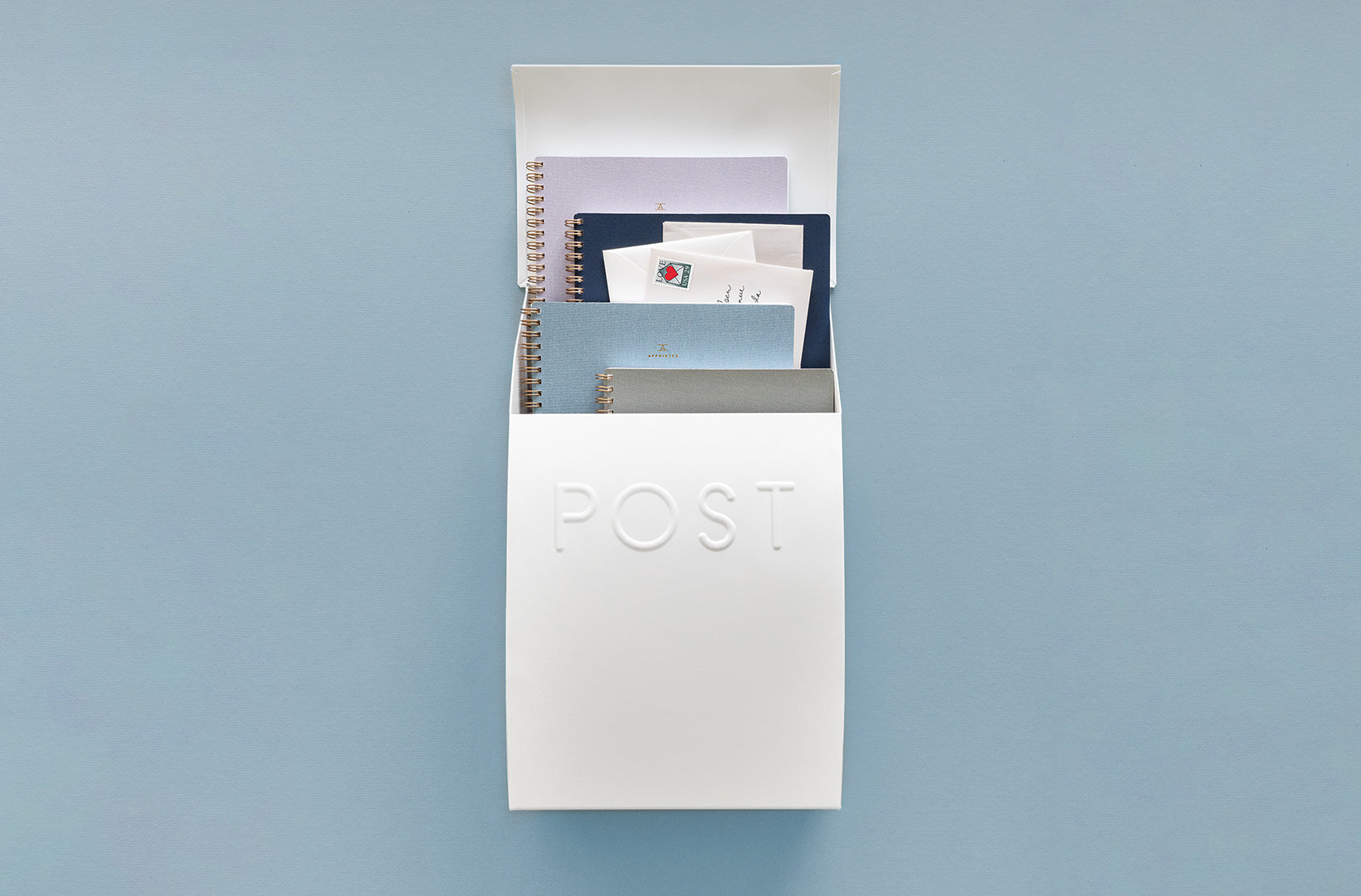 A white mailbox hinges open at the lid, revealing copious letters and notebooks inside. The mailbox is vertical and sits against a blue-gray wall.