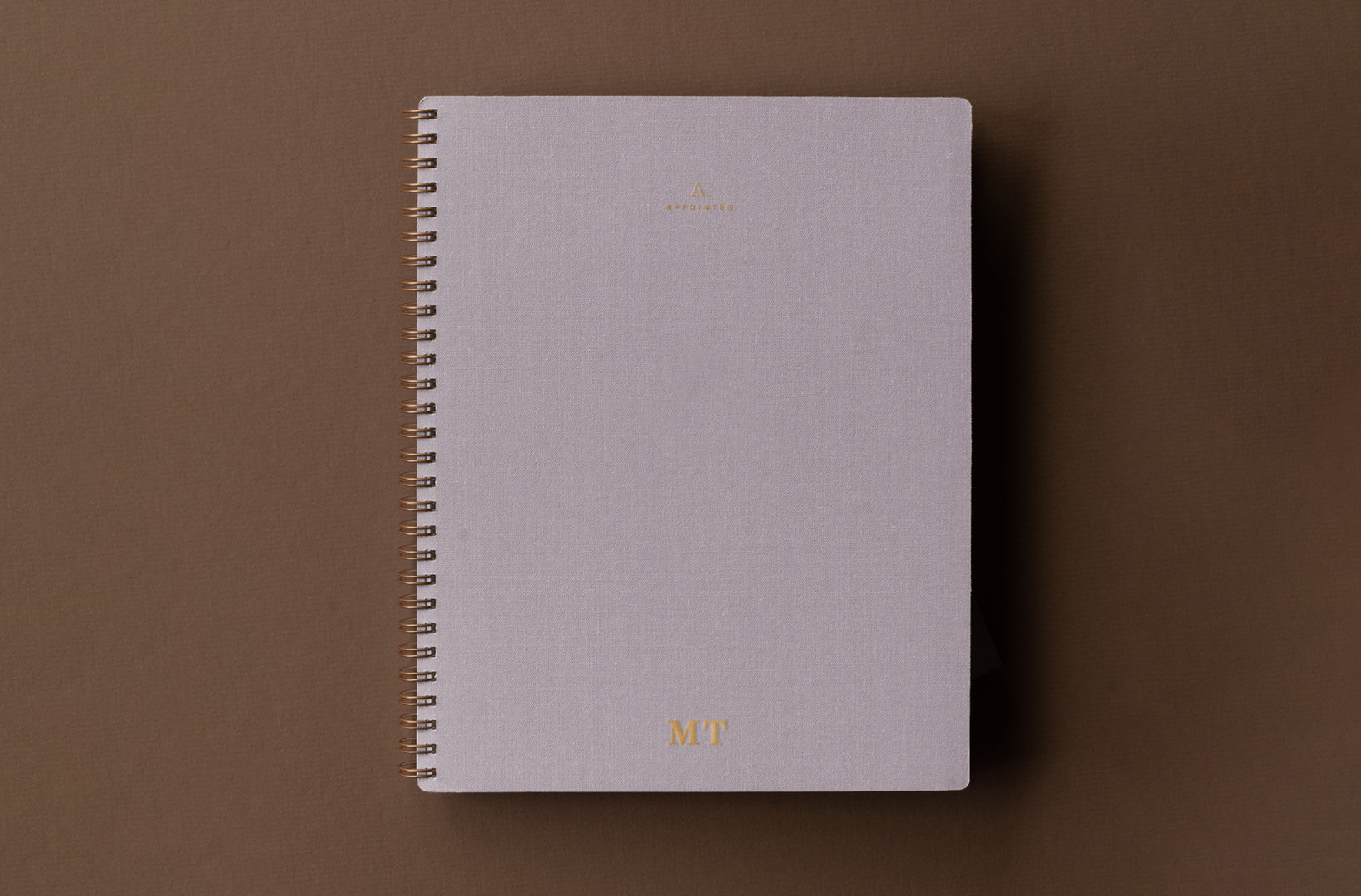 A lavender gray notebook with wire-o binding and gold foil monogram sits against a brown background