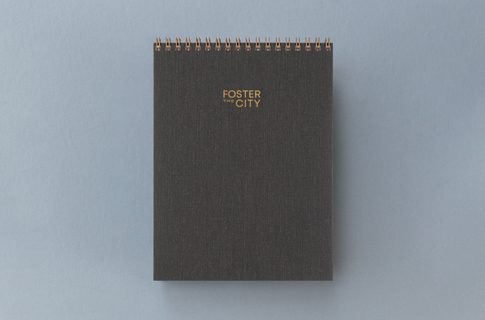 A charcoal gray office notepad with wire-o binding and foil stamped custom logo sits against a gray-blue background