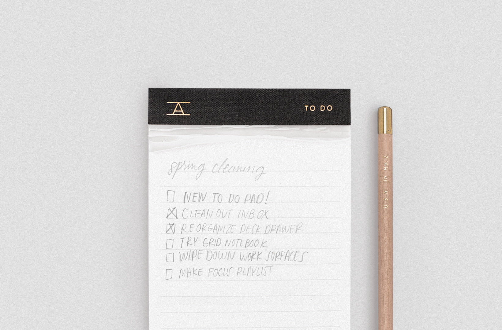 An Appointed To Do Pad with charcoal bookcloth binding and gold foil details sits against a gray back ground with a natural no. 2 pencil with gold cap sitting next to it. There is a to-do list scrawled in pencil on the pad and you can see the remnants of previous torn pages