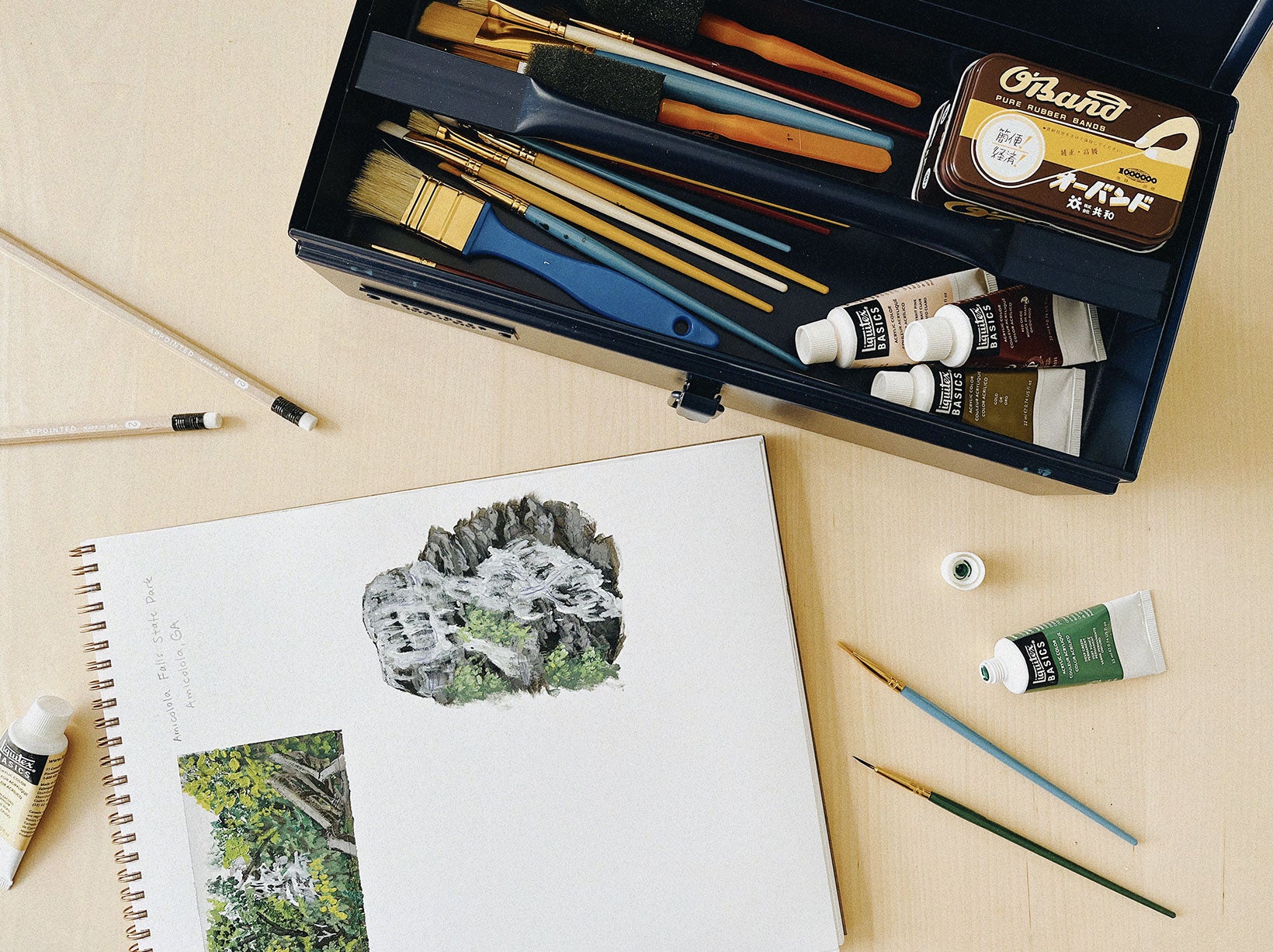 the toolbox in midnight is filled with art supplies including paints, pencils, and brushes. a sketch pad sits open with paintings of natural scenes against a wood table.