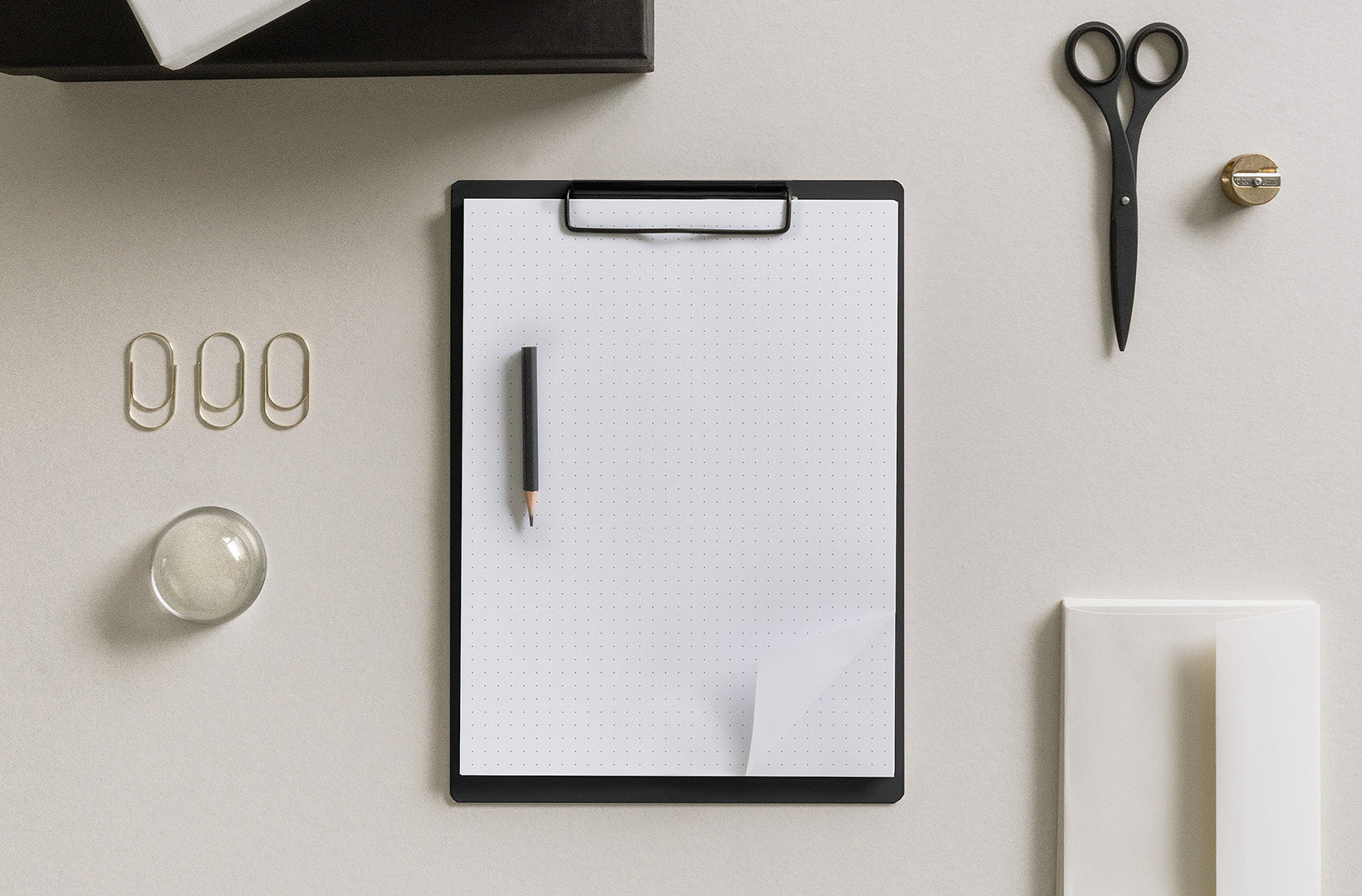 The Writing clipboard with Dot Grid Pad sits on a beige background. Other accessoreis are strewn about, including scissors, paper clips, envelopes, and a brass sharpener