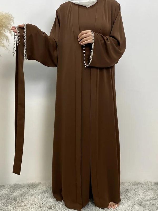 Femme portant une abaya marron avec des broderies blanches aux manches, tenant une ceinture assortie dans une main, portant un hijab blanc, posant sur un fond neutre avec un vase de pampas à gauche