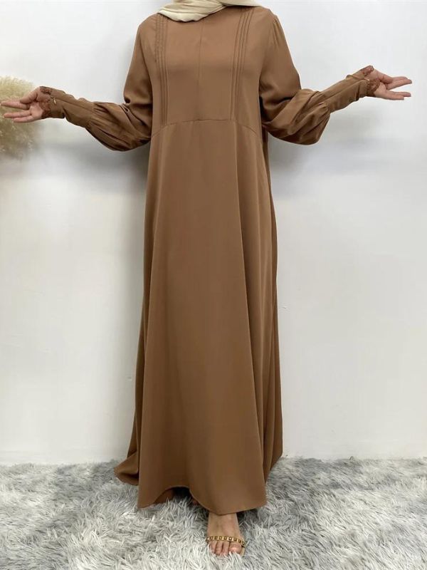 La femme porte une abaya marron clair qui est longue et fluide, tombant jusqu'au sol. Elle a les bras légèrement écartés, ce qui permet de voir les manches amples de la robe. La tenue est simple et élégante, avec des plis discrets sur le haut qui ajoutent de la texture à la robe. Le hijab beige clair complète l'ensemble en apportant une touche de douceur. Cette abaya est à la fois modeste et sophistiquée, parfaite pour un look élégant tout en restant confortable