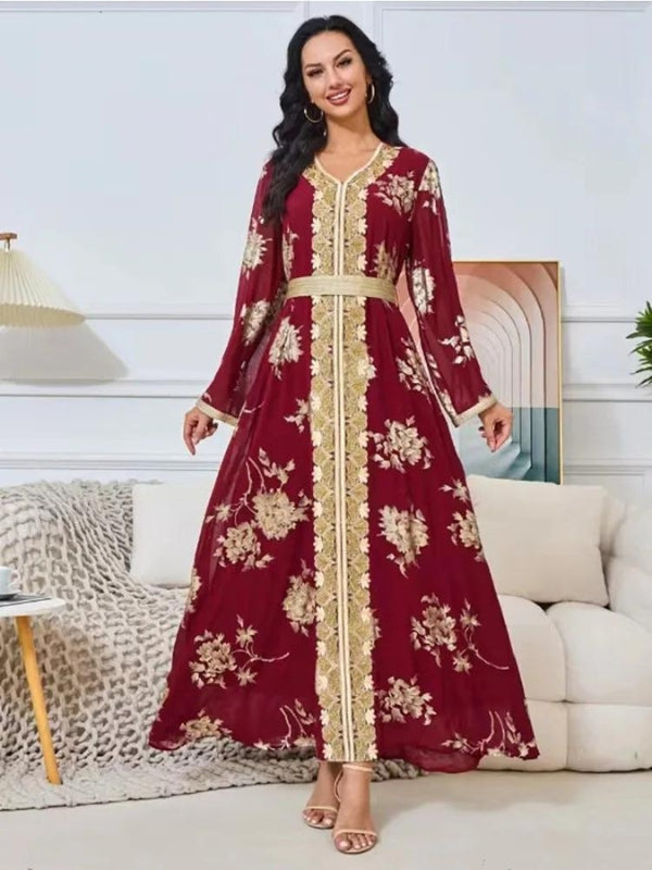 La femme sur cette image porte un magnifique caftan long de couleur bordeaux avec des motifs floraux dorés. Le caftan est orné d'une bande brodée dorée qui descend au centre, mettant en valeur la coupe en V du col. Elle porte une ceinture assortie à la taille, ce qui accentue sa silhouette. Ses cheveux noirs et bouclés tombent sur ses épaules, et elle arbore des boucles d'oreilles créoles dorées, complétant son look élégant. Elle est debout dans une pièce bien éclairée, devant un canapé clair, ce qui met en valeur la beauté et l'élégance de sa tenue.