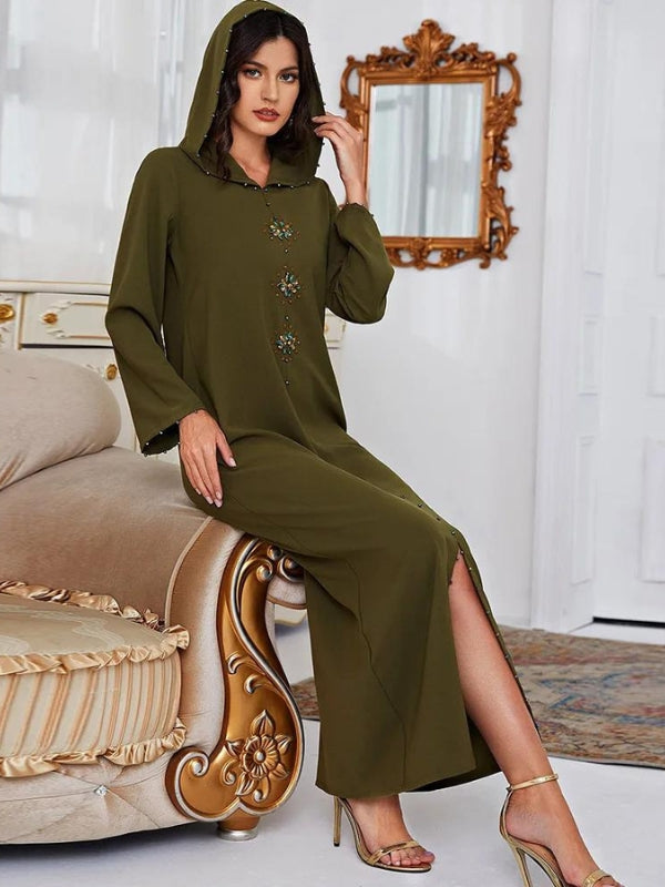 Une femme est assise sur un canapé luxueux, portant une djellaba de couleur kaki avec une capuche. La djellaba est ornée de broderies délicates sur le devant, ajoutant une touche d'élégance à la tenue. Les manches longues et le tissu fluide offrent un confort optimal tout en restant sophistiqués. La femme porte des talons hauts dorés, accentuant son allure élégante. L'arrière-plan de la pièce, avec un miroir encadré de bois doré et un canapé aux détails opulents, complète l'ambiance chic et raffinée de la scène.