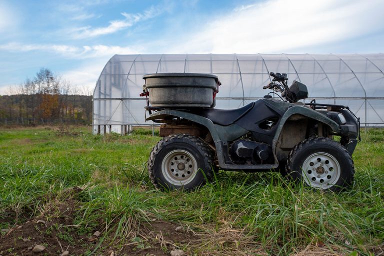 Beautiful agricultural scene with atv off road farm work vehicle with a bin strapped to the back and a green house in the background under a blue sky with white clouds. No people, shot in natural