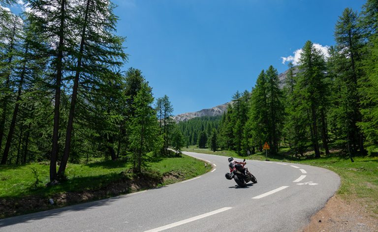 Unrecognizable tourist on a motorcycle leans in to ride into an empty turn in the sunlit forest in the French Alps. Traveler on motorbike having fun on a summer road trip along Route des Grandes Alpes