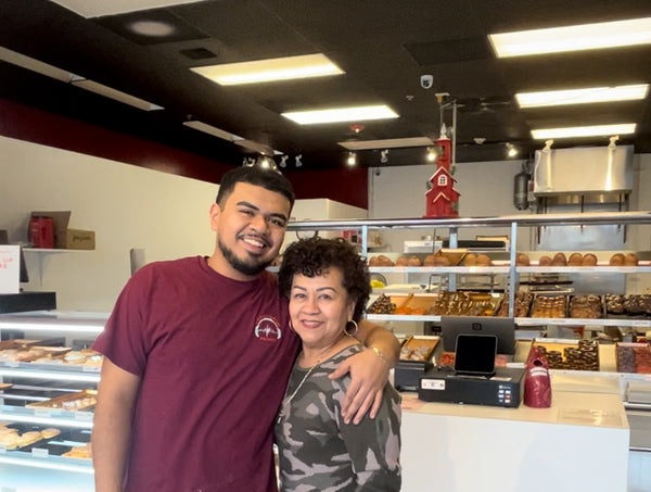 A Mexican man with facial hair stands with his arm around his mom in a donut shop