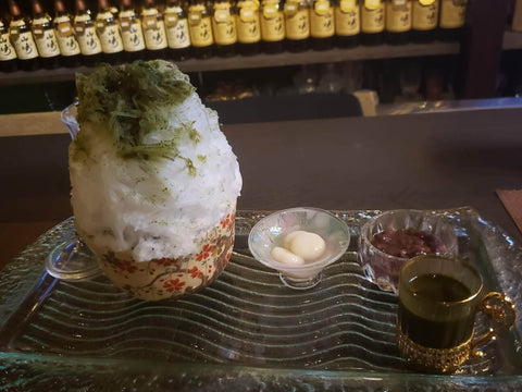 A bowl of Japanese shaved ice with a green tea syrup next to mochi and red bean paste