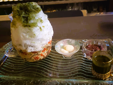 A bowl of fluffy shaved ice with green tea topping next to a bowl of mochi and red beans