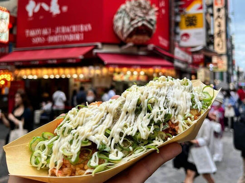 A hand holds a tray of takoyaki in front of a popular Osaka shop