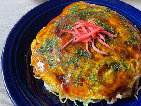 A plate of thinner Hiroshima okonomiyaki with a thick sauce and ginger on top sits on a table