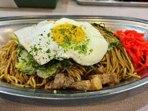 A plate of yakisoba noodles topped with an egg, pork and pickled ginger