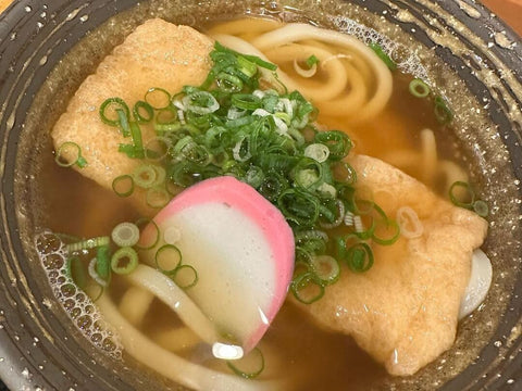 A bowl of kitsune udon with a strip of fried tofu, lots of green onion and some kamaboko