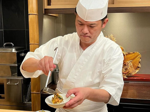 A Japanese chef pours something over a dish with a metal ladle