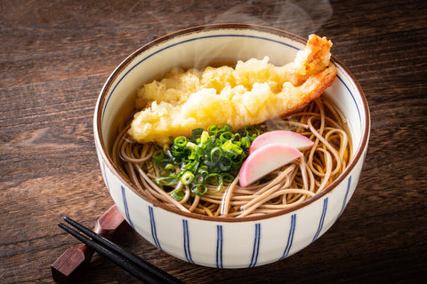 A bowl of soba with a clear broth, kamaboko, green onion and tempura on top