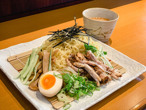 A plate with cold noodles, meat, egg and veggies over bamboo next to a cup of broth