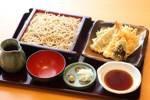 A bamboo box of cold soba noodles next to tempura, tsuyu dipping sauce, and other add-ins