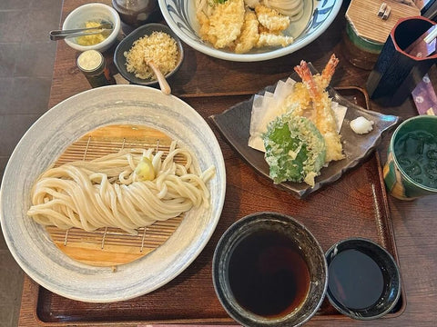 A plate of a bundle of cold udon next to a bowl of dipping soup and tempura