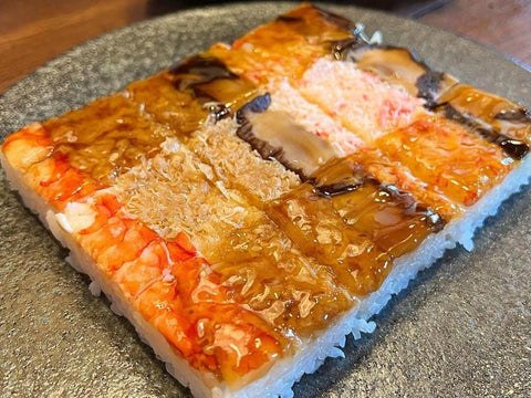 An arrangement of square hakozushi sushi of various colors, fish types, and sauce toppings