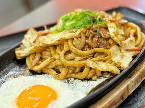 A hot skillet with a stir-fried yaki-udon with meat and vegetables and a fried egg on the side