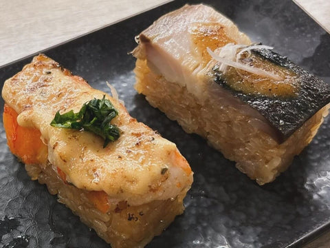 Two pieces of hakozushi, one with salmon and cheese and one with mackerel and sauce