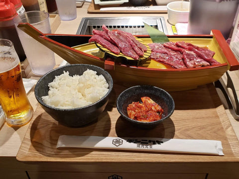 Meat sits on a boat platter with a bowl of rice and kimchi in front of a grill