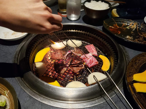 A pair of tongs held by a hand goes to flip meat on a yakiniku grill