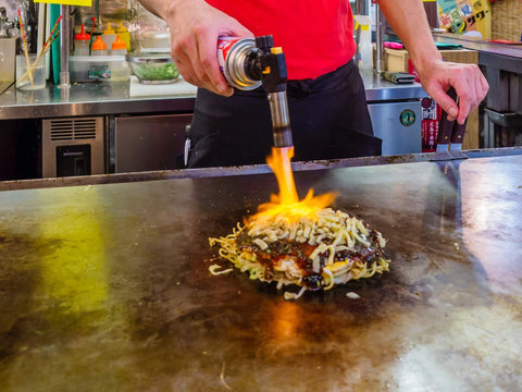 A hand uses a blow torch to melt cheese on top of a serving of okonomiyaki