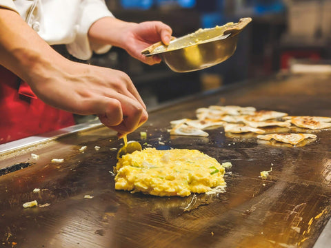 A hand spoons okonomiyaki batter carefully onto a flat grill