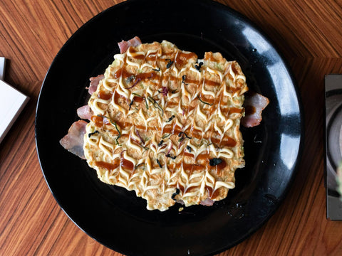 One okonomiyaki sits on a plate over bacon and with mayo and sauce on top